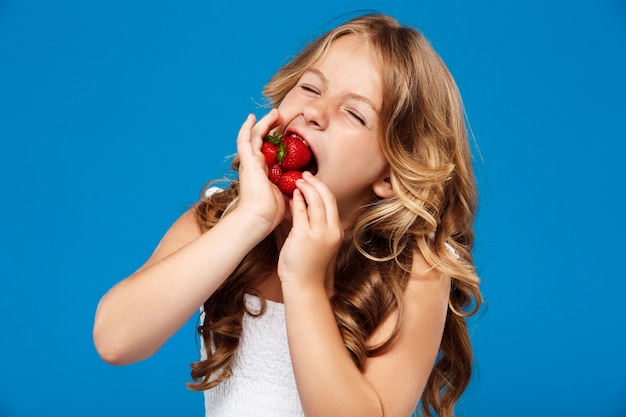 Jong mooi meisje dat aardbei over blauwe muur eet