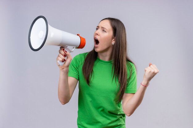 Jong mooi meisje dat een groene t-shirt draagt die aan emotionele en ongeruste megafoon schreeuwt die zich over geïsoleerde witte achtergrond bevindt