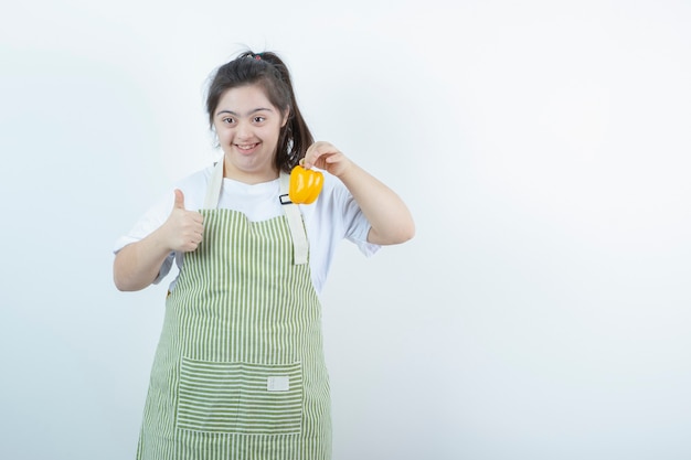 Jong mooi meisje dat in geruite schort gele paprika houdt en duim toont.
