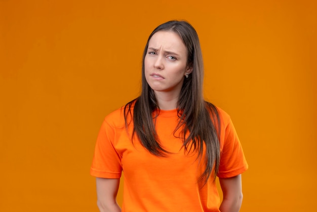 Jong mooi meisje dat oranje t-shirt draagt dat camera bekijkt ontevreden met droevige uitdrukking op gezicht dat zich over geïsoleerde oranje achtergrond bevindt