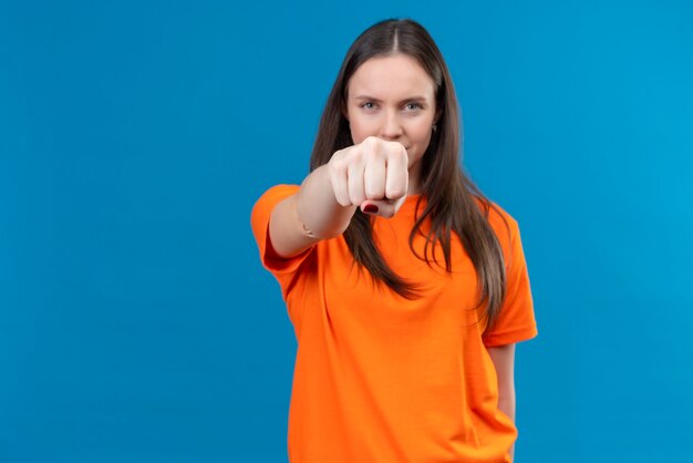 Jong mooi meisje dat oranje t-shirt draagt dat met vuist naar camera richt die zich zelfverzekerd over geïsoleerde blauwe achtergrond bevinden