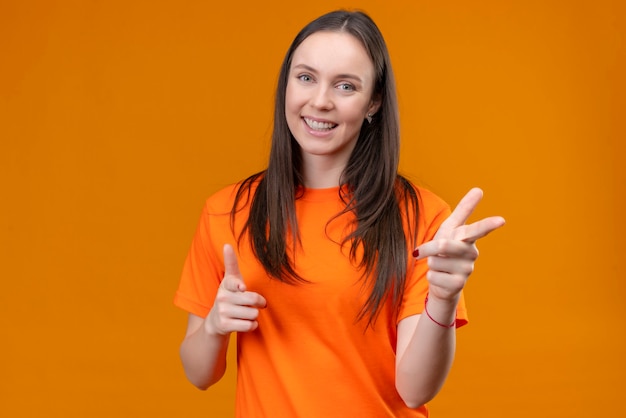 Jong mooi meisje dat oranje t-shirt draagt dat naar camera richt die vrolijk positief en gelukkig glimlacht over geïsoleerde oranje achtergrond