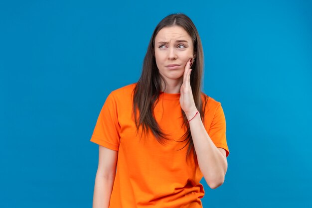 Jong mooi meisje dat oranje t-shirt draagt dat onwel kijkt wat betreft haar wang voelt kiespijn die zich over geïsoleerde blauwe achtergrond bevindt