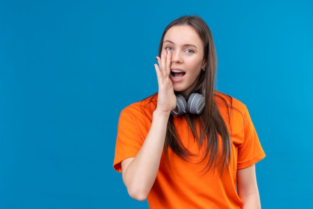 Jong mooi meisje dat oranje t-shirt met hoofdtelefoons draagt die een geheim met hand dichtbij mond vertelt gelukkig en positief status over geïsoleerde blauwe achtergrond