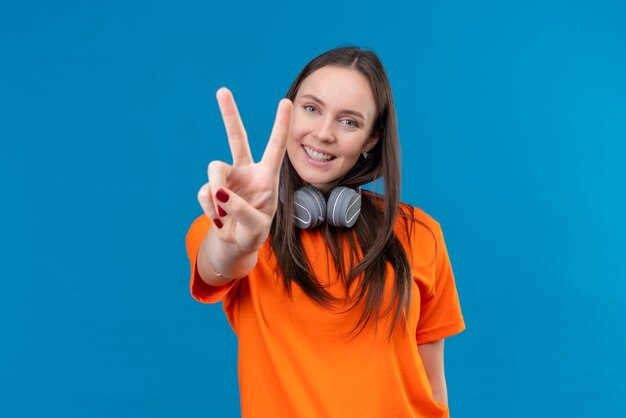 Jong mooi meisje dat oranje t-shirt met hoofdtelefoons draagt die vrolijk glimlacht toont overwinningsteken dat zich over geïsoleerde blauwe achtergrond bevindt