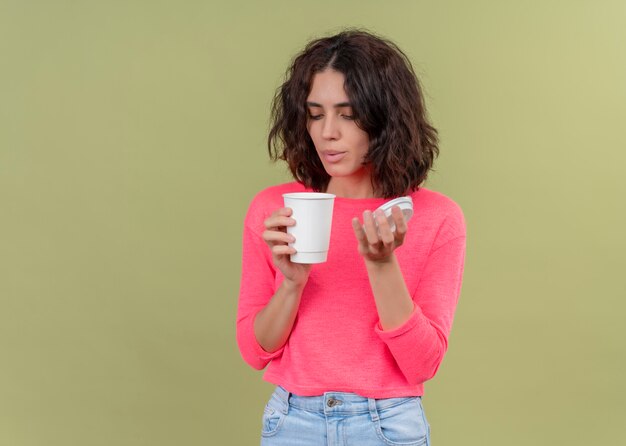 Jong mooi meisje dat plastic koffiekop houdt en ernaar blaast op geïsoleerde groene achtergrond met exemplaarruimte