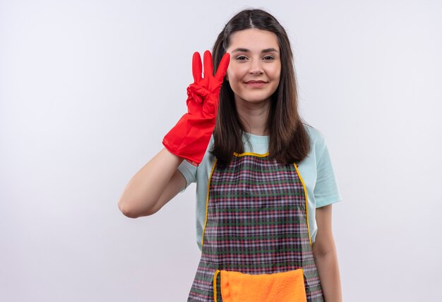 Jong mooi meisje dat schort en rubberhandschoenen draagt die en met vingers nummer drie glimlachen benadrukken