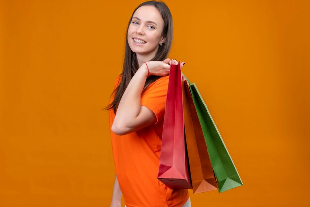 Jong mooi meisje die een oranje t-shirt dragen die papieren zakken houdt die camera bekijken die vrolijk glimlacht over geïsoleerde oranje achtergrond bevindt