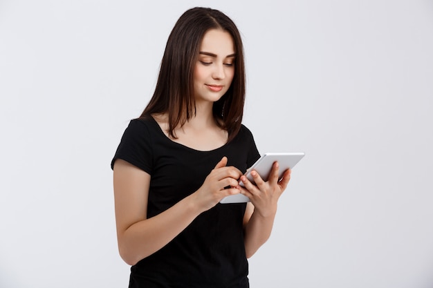 Jong mooi meisje in de zwarte tablet van de t-shirtholding en het bekijken met belangstelling het scherm over witte muur