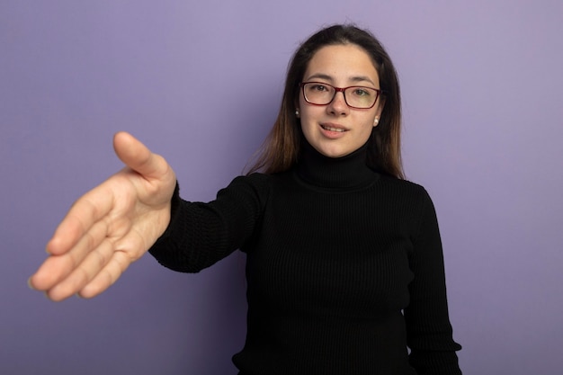 Jong mooi meisje in een zwarte coltrui en glazen die hand het glimlachen vriendelijke groet aanbieden