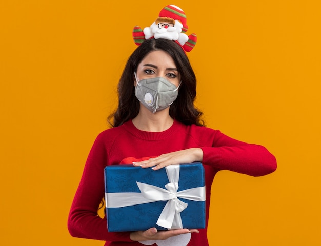 Jong mooi meisje met de hoofdband en trui van de kerstman met een beschermend masker met een cadeaupakket geïsoleerd op een oranje muur