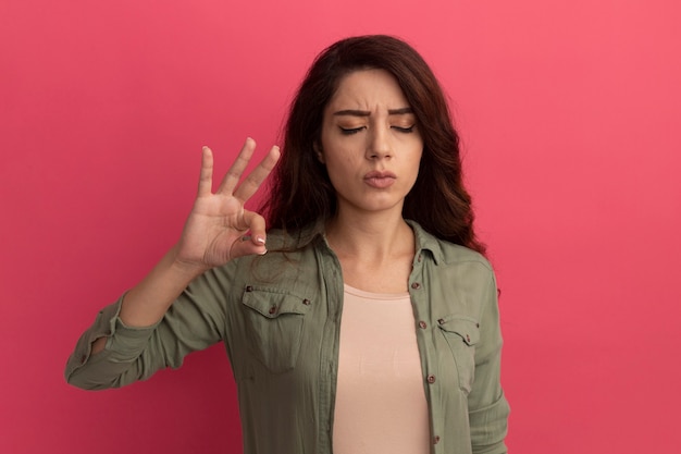 Jong mooi meisje met gesloten ogen die olijfgroen t-shirt dragen die ok gebaar tonen dat op roze muur wordt geïsoleerd