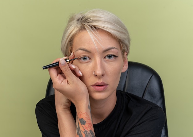 Gratis foto jong mooi meisje teken pijl met eyeliner geïsoleerd op olijfgroene muur