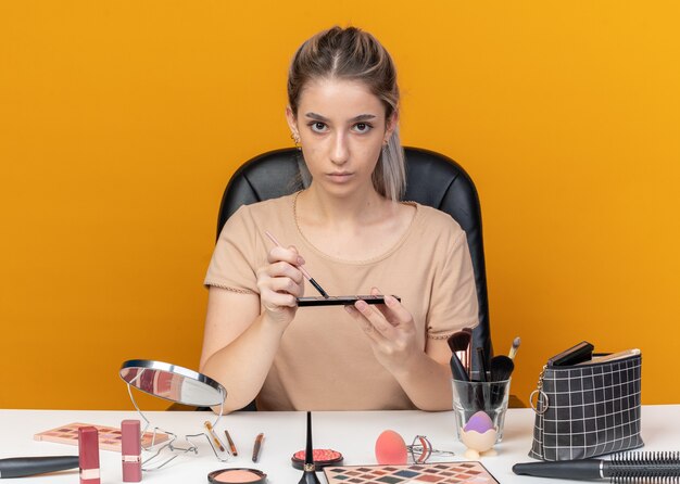 jong mooi meisje zit aan tafel met make-up tools oogschaduw toe te passen met make-up borstel geïsoleerd op oranje muur