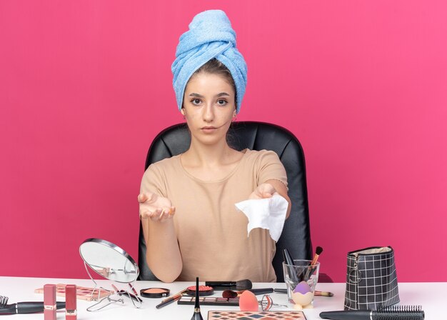 jong mooi meisje zit aan tafel met make-uptools gewikkeld haar in een handdoek en steekt servet uit dat op roze muur is geïsoleerd