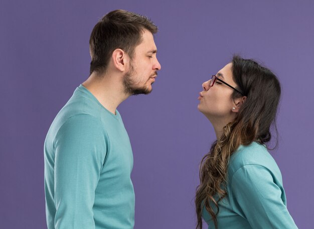 Jong mooi stel in blauwe vrijetijdskleding, gelukkige en vrolijke man en vrouw die gelukkig verliefd gaan kussen over de paarse muur