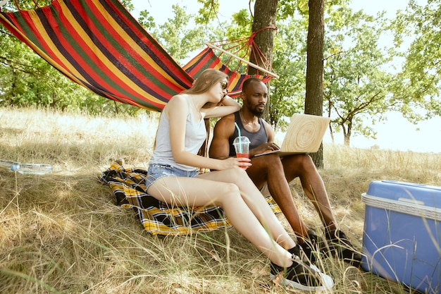 Jong multi-etnisch internationaal romantisch koppel buiten op de weide in zonnige zomerdag. Afro-Amerikaanse man en blanke vrouw met picknick samen. Concept van relatie, zomer.