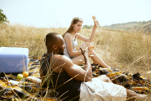 Jong multi-etnisch internationaal romantisch koppel buiten op de weide in zonnige zomerdag. Afro-Amerikaanse man en blanke vrouw met picknick samen. Concept van relatie, zomer.