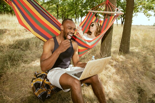 Jong multi-etnisch internationaal romantisch koppel buiten op de weide in zonnige zomerdag. Afro-Amerikaanse man en blanke vrouw met picknick samen. Concept van relatie, zomer.