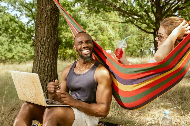 Jong multi-etnisch internationaal romantisch koppel buiten op de weide in zonnige zomerdag. Afro-Amerikaanse man en blanke vrouw met picknick samen. Concept van relatie, zomer.