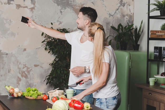 Jong paar dat selfie op mobiele telefoon neemt terwijl het snijden van groenten