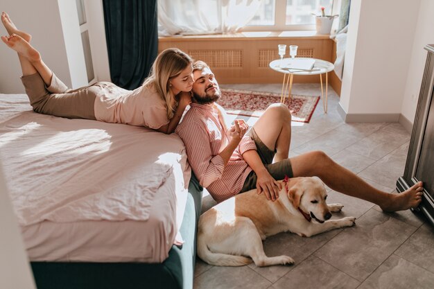 Jong paar liefhebbers rusten in de slaapkamer. Meisje in comfortabele kleren ligt op bed en kijkt naar haar vriendje terwijl hij Labrador aait.