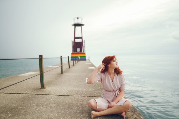 jong roodharig meisje in een grote ronde hoed en roze jurk zittend op de ligplaats in de buurt van de oceaan