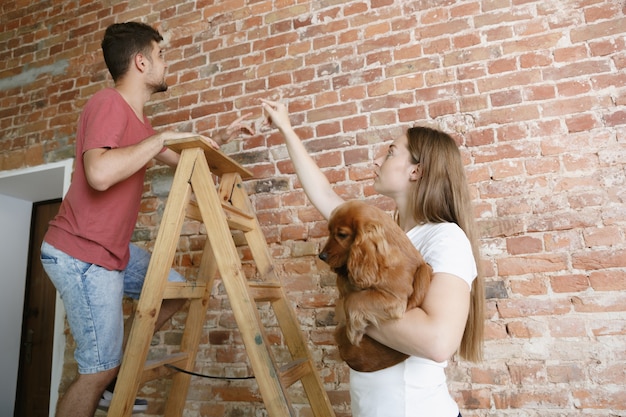 Jong stel dat zelf appartementreparatie doet. Getrouwde man en vrouw die huis make-over of renovatie doen. Concept van relaties, familie, huisdier, liefde. Bespreek het toekomstige ontwerp aan de muur.