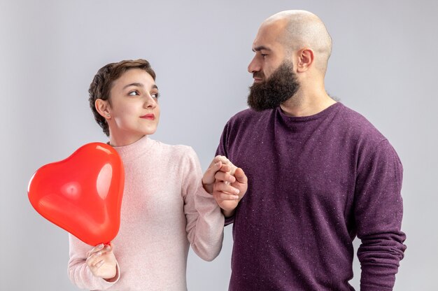 jong stel in vrijetijdskleding bebaarde man kijken naar zijn gelukkige vriendin met kort haar hartvormige ballon vasthouden gelukkig verliefd Valentijnsdag vieren staande over witte muur