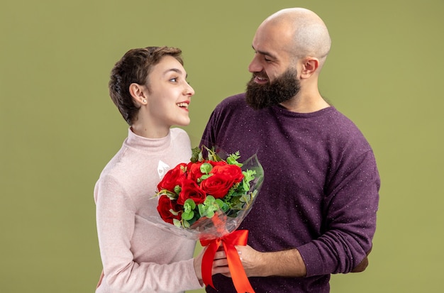 jong stel in vrijetijdskleding gelukkig bebaarde man die een boeket rode rozen geeft aan zijn lachende vriendin Valentijnsdag vieren staande over groene muur