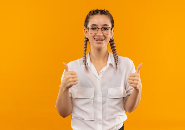 Jong studentenmeisje dat in glazen met vlechten in wit overhemd naar de voorzijde kijkt die vrolijk glimlachend duimen toont die zich over oranje muur bevinden