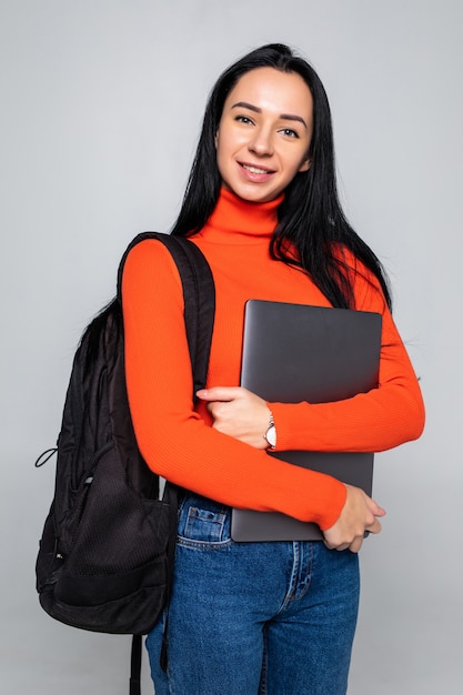 Jong studentenmeisje dat op grijze muur wordt geïsoleerd, die bij camera glimlacht, laptop tegen borst drukt, rugzak draagt, klaar om naar studies te gaan, nieuw project begint en nieuwe ideeën voorstelt.