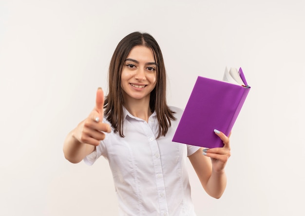 Jong studentenmeisje die in wit overhemd open boek houden die duimen die omhoog glimlachen status over witte muur tonen