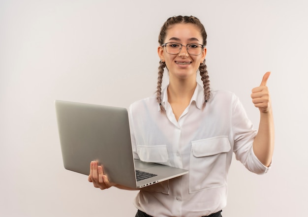 Jong studentenmeisje in glazen met vlechten in wit overhemd die laptop houden die naar de voorkant glimlachen glimlachend vrolijk tonen duimen omhoog die zich over witte muur bevinden