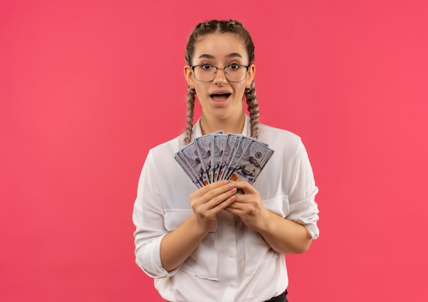 Jong studentenmeisje in glazen met vlechtjes in wit overhemd die contant geld tonen dat verbaasd en verbaasd over roze muur kijkt