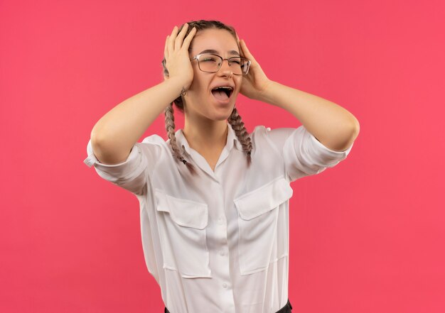 Jong studentenmeisje in glazen met vlechtjes in wit overhemd die opzij kijken met haar hoofd met handen schreeuwen in paniek die zich over roze muur bevinden