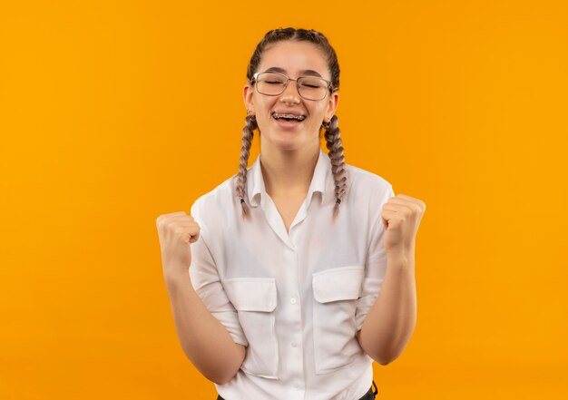 Jong studentenmeisje in glazen met vlechtjes in wit overhemd die vuisten balde blij en opgewonden verheugend zich over haar succes dat zich over oranje muur bevindt