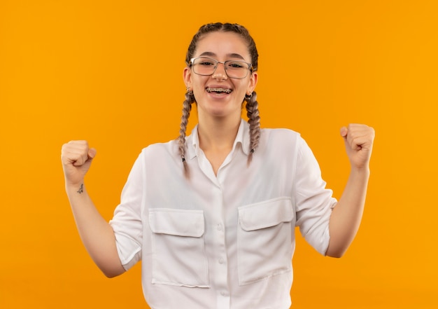 Jong studentenmeisje in glazen met vlechtjes in wit overhemd die vuisten balde blij en opgewonden verheugend zich over haar succes dat zich over oranje muur bevindt