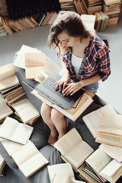 Jong tienermeisje dat de laptop computer met behulp van die door vele boeken wordt omringd.