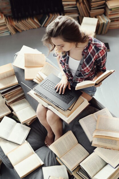 Jong tienermeisje dat de laptop computer met behulp van die door vele boeken wordt omringd.