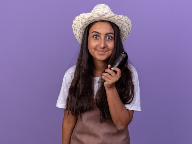 Jong tuinmanmeisje in schort en de zomerhoed die aubergine het gelukkige en positieve glimlachen houden die zich over purpere muur bevinden