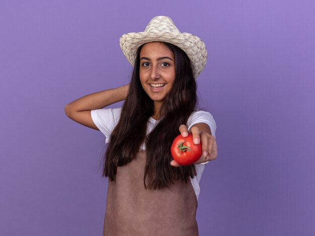 Jong tuinmanmeisje in schort en de zomerhoed die verse tomaten met glimlach op gezicht houden die zich over purpere muur bevinden