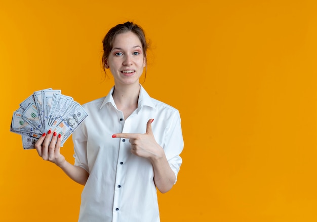 Jong verrast blond russisch meisje houdt en wijst op geld op oranje met exemplaarruimte