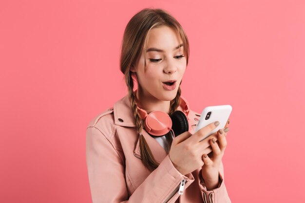 Jong verrast mooi meisje met twee vlechten in leren jas met koptelefoon die verbaasd naar mobiel kijkt terwijl ze tijd doorbrengt op roze achtergrond