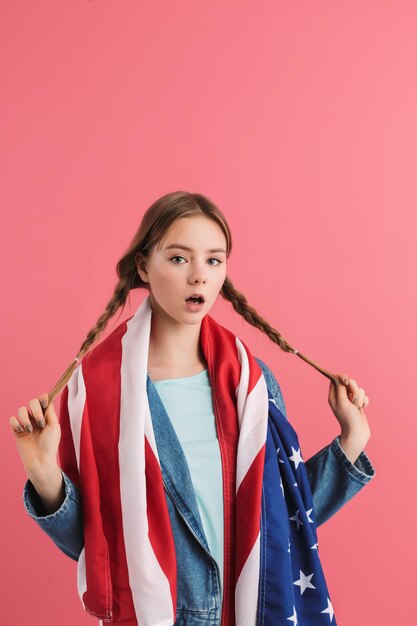 Jong verrast tienermeisje met twee vlechten die verbaasd in de camera kijken met Amerikaanse vlag op schouders over roze geïsoleerde achtergrond