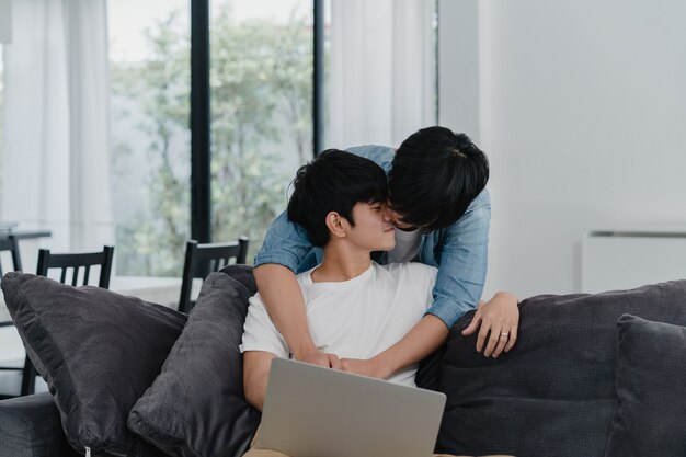 Jong Vrolijk paar die computerlaptop met behulp van bij modern huis. Aziatische LGBTQ + mannen gelukkig ontspannen plezier met behulp van technologie kijken naar film op internet samen terwijl liggend bank in de woonkamer in huis.