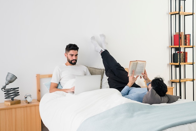 Jong vrolijk paar die op bed liggen die het boek lezen en mobiele telefoon met behulp van