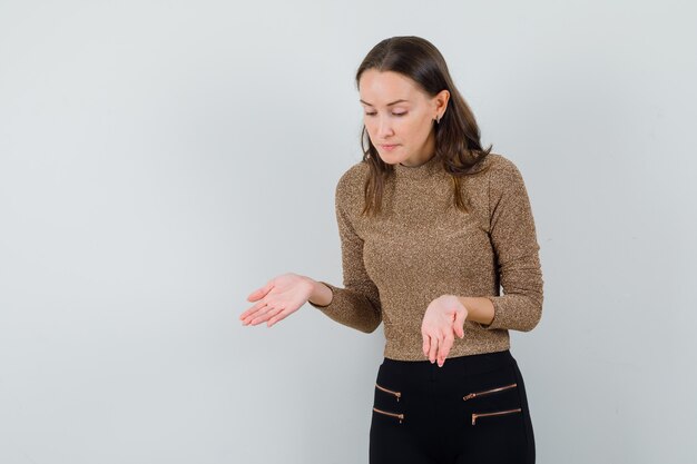 Jong wijfje dat iets toont bij vloer met open handpalmen in gouden blouse en ernstig kijkt. vooraanzicht.