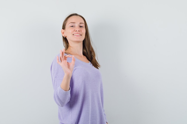 Jong wijfje dat ok gebaar in lila blouse toont en tevreden kijkt. vooraanzicht.