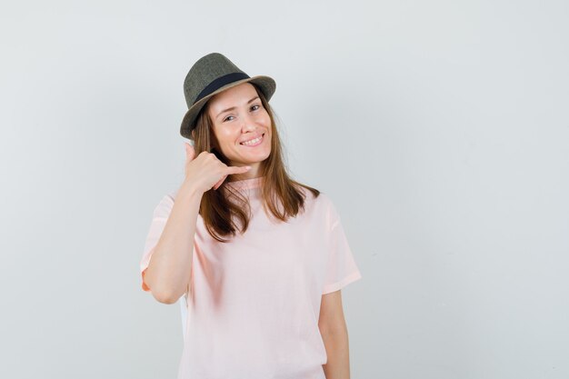 Jong wijfje dat telefoongebaar in roze t-shirt, hoed toont en nuttig, vooraanzicht kijkt.